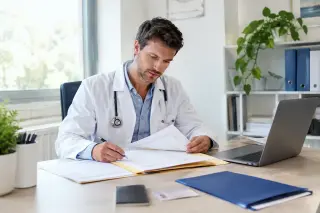 Médecin en blouse blanche, assis à un bureau dans un cabinet moderne, entouré de dossiers et de documents officiels, préparant son installation professionnelle dans une ambiance organisée et lumineuse.